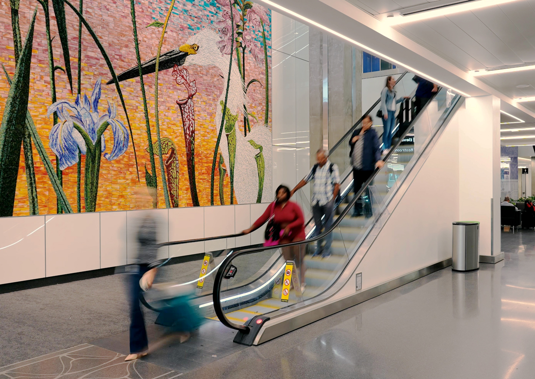 Passengers ride an escalator at Tampa International Airport’s Express Curbsides, passing a large mosaic artwork integrated into the interior circulation space.