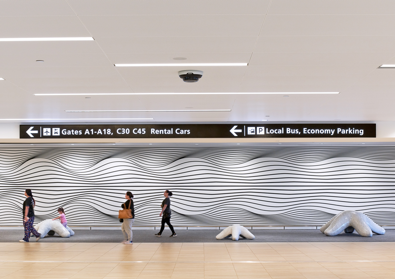 Passengers walk through a Tampa International Airport corridor featuring directional signage and a large wall installation with wave-like graphics and sculptural seating.