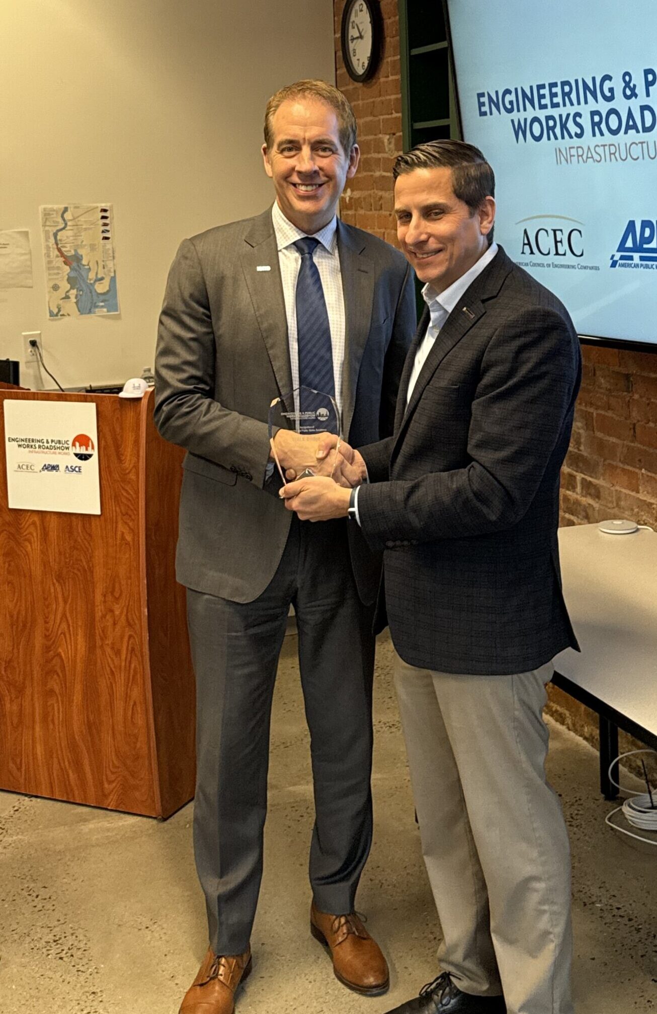 Two men standing next to one another shaking hands and holding an award.