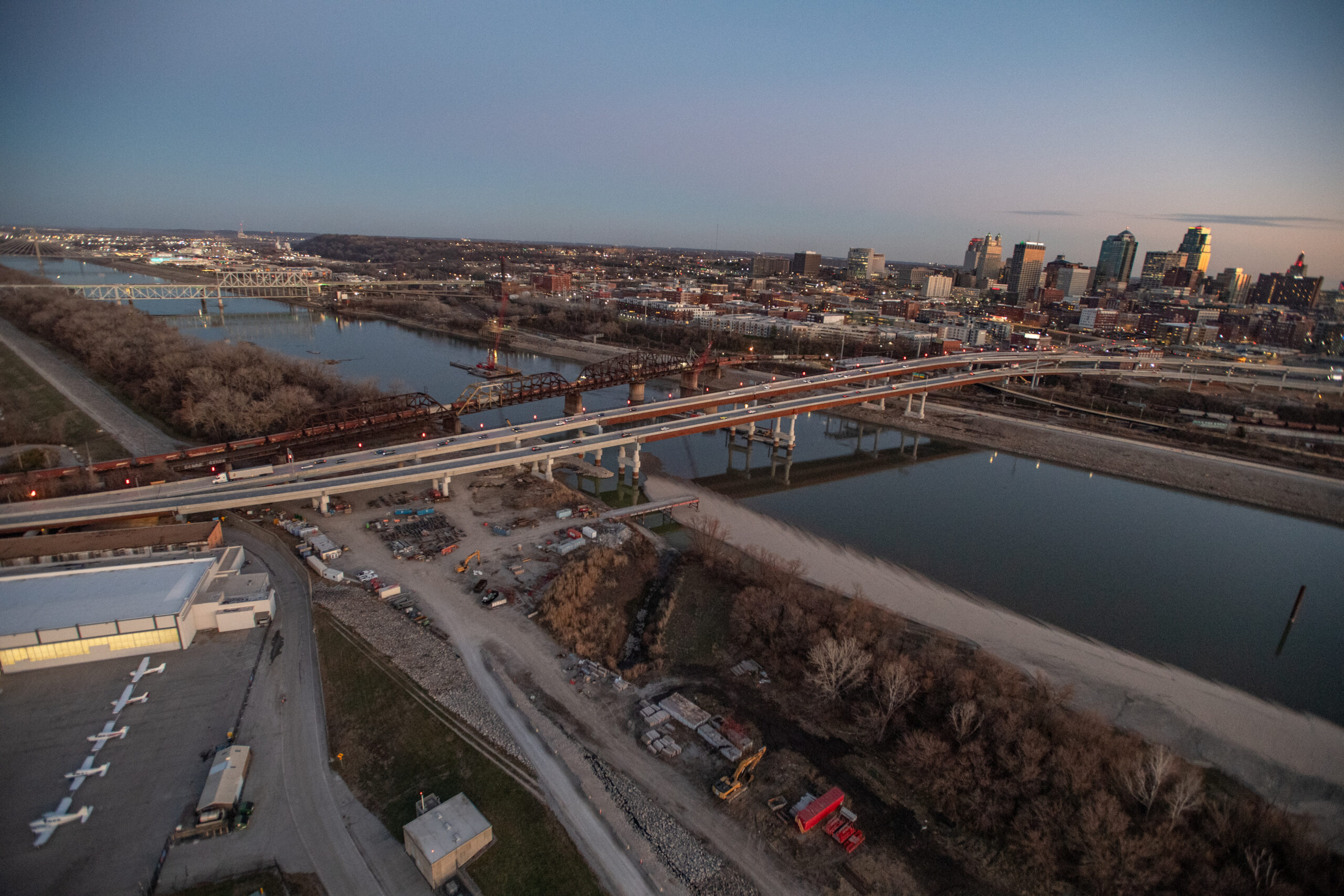 An aerial view of the Buck O’Neil Bridge project showing multiple bridge spans under construction across the river with cranes, equipment and Kansas City’s skyline in the distance.