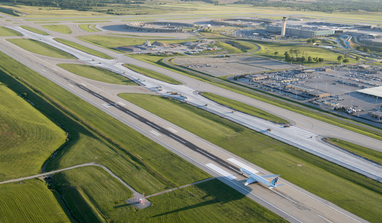 Aerial view of an active airport airfield showing a runway, taxiways, and a taxiing aircraft, with terminal buildings and control tower in the background, illustrating large-scale aviation infrastructure and coordinated capital development.