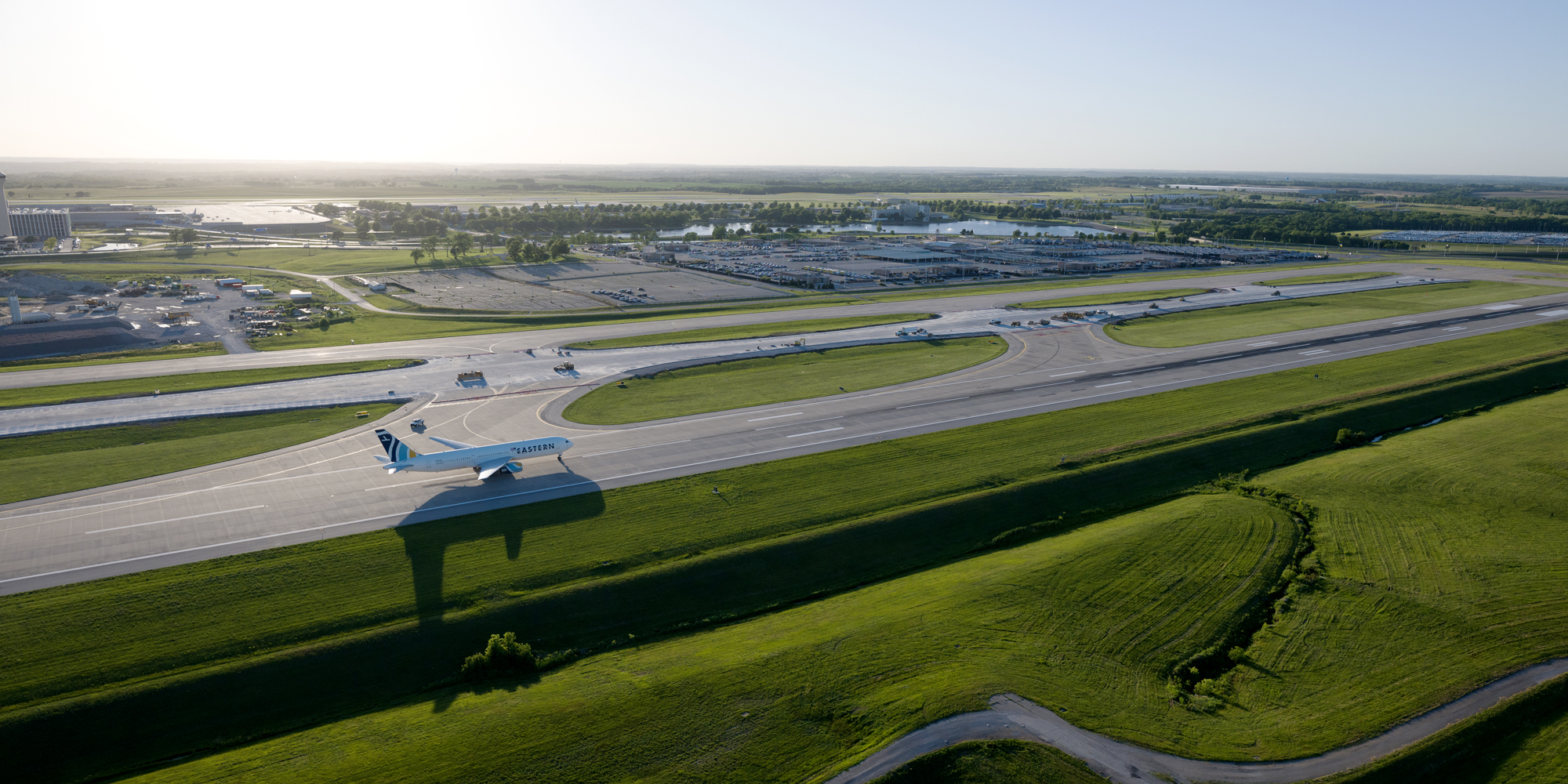 Aerial view of an active airport airfield showing a runway, taxiways, and a taxiing aircraft, with terminal buildings and control tower in the background, illustrating large-scale aviation infrastructure and coordinated capital development.